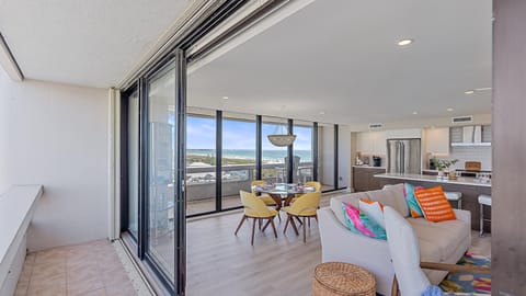 Modern open-plan living area with a kitchen, dining table, and seating area featuring colorful cushions. Sliding glass doors open to a balcony with an ocean view.