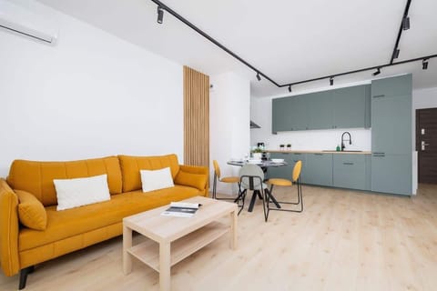A modern living room with mustard yellow sofas, a light wooden coffee table, and a minimalist design, featuring pale green kitchen cabinets in the background.

