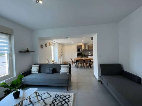 "This open plan living room features grey tiled floor that creates a modern, easy to maintain base. In the heart of the space, a comfortable sofa coexists with a sofa bed, offering versatility and space for guests. The living area opens harmoniously to the kitchen, creating a friendly and welcoming atmosphere."