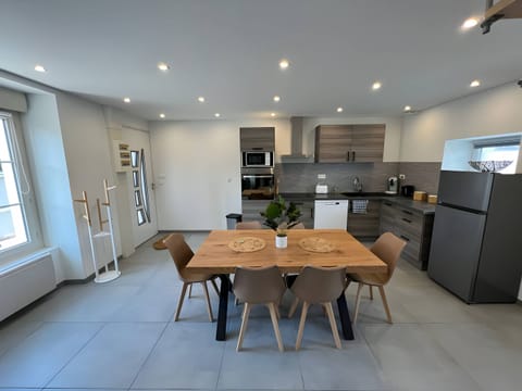 "This fitted and fully equipped kitchen is a friendly and functional space. The grey tiled floor creates an elegant contrast with the white walls, bringing a touch of modernity. In the center of the kitchen, a large wooden table seats up to six people on comfortable brown chairs."