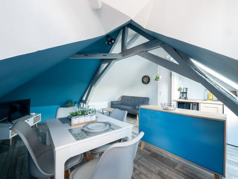 "This room is a living space under the attic, combining a dining room, a kitchen and a living room. Gray beams on the ceiling add rustic charm. A blue wall creates a focal point, and a TV area is dedicated to entertainment. The set offers a comfortable and elegant setting to relax and share meals."