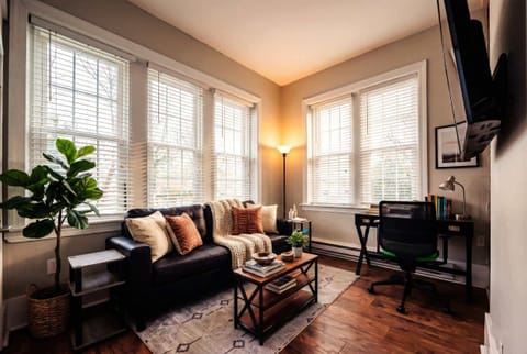Bright living area with sofa seating, wall-mounted TV, desk, and large windows.