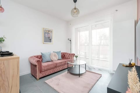 A bright and cozy living room with a pink sofa, light-colored furniture, and large windows allowing plenty of natural light.

