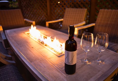 This outdoor space is a perfect evening setting, featuring a rustic wooden table with an integrated fire pit, accompanied by a bottle of wine and glasses.