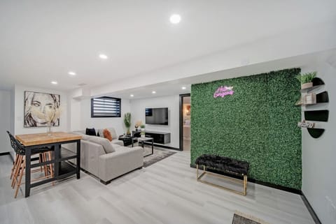 Spacious modern living area with comfortable sectional sofa & large TV. Features a unique green selfie wall, stylish high-top table, and vibrant artwork. Perfect for relaxing or social gatherings. Contemporary design & inviting ambiance