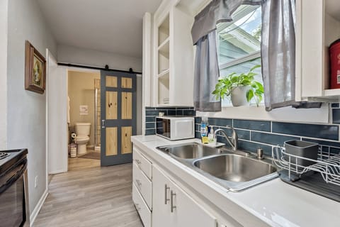 Cute and compact kitchen with open shelving, a microwave, and big window for natural light—plus a peek into the full bath.