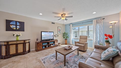 A living room with a TV, a ceiling fan, a sofa, a recliner, and a coffee table on a rug. There's a cabinet with a vase, a plant, a floor lamp, red flowers, and a large window with curtains.