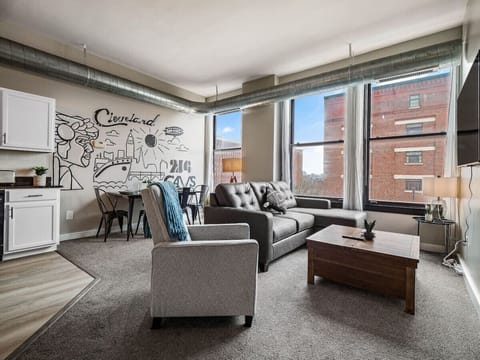 -Bright living room with large industrial windows bringing in natural light - Cozy sofa and armchair perfect for streaming or relaxing after exploring Cleveland - Open concept layout with dining table and Cleveland wall art accent