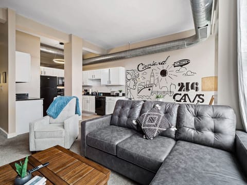 Stylish living space with sectional sofa, accent chair, and wood coffee table - Large wall mural showcasing Cleveland pride with modern artistic design - Open layout connects seamlessly with kitchen and dining area for comfort