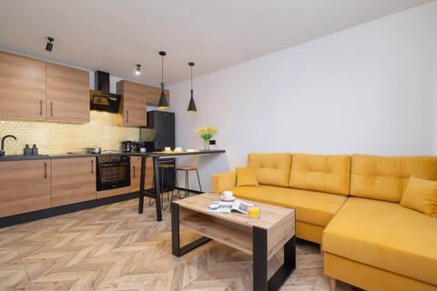 A modern living room with a warm yellow sofa, wooden furniture, and a sleek black kitchen, complemented by stylish lighting.
