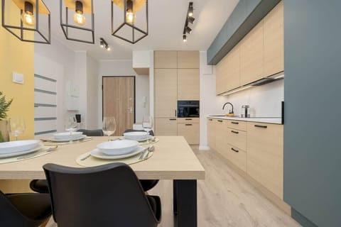 Dining area featuring a light wooden table, black chairs, and a yellow wall in the background, with modern lighting fixtures.