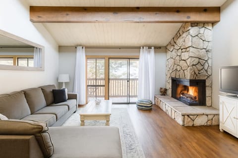 Cozy living room featuring a large, comfortable sofa, high ceilings, and a beautiful stone fireplace—perfect for relaxing after a day on the mountain. Step out onto the private balcony and enjoy incredible views where you can actually watch skiers coming 