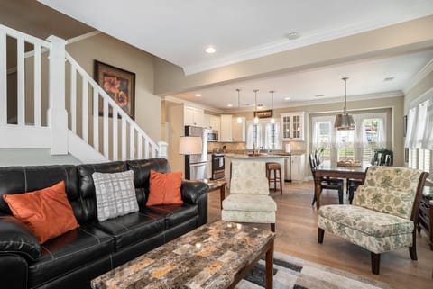 Features a black leather sofa with orange and grey throw pillows, a marble-topped coffee table, and two patterned armchairs. The space seamlessly transitions into a kitchen and dining area.