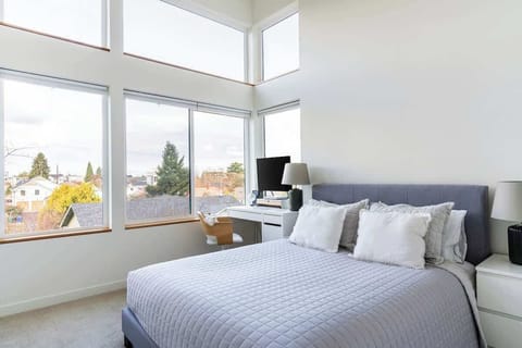 Light-filled modern bedroom with expansive windows, plush bedding, and a sleek workspace overlooking scenic neighborhood views.
