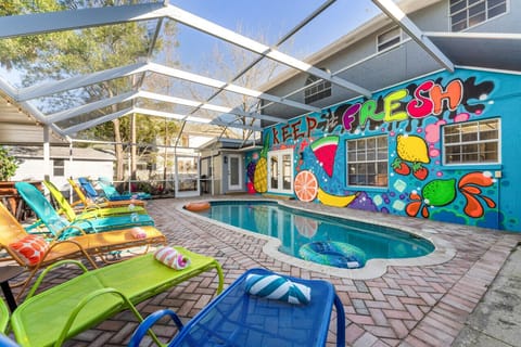 Featuring a stunning outdoor scene, with a large swimming pool as the focal point. The pool is surrounded by colorful chairs and sun loungers, creating an inviting and relaxing atmosphere. The vibrant mural painted on its side adds an artistic touch to th