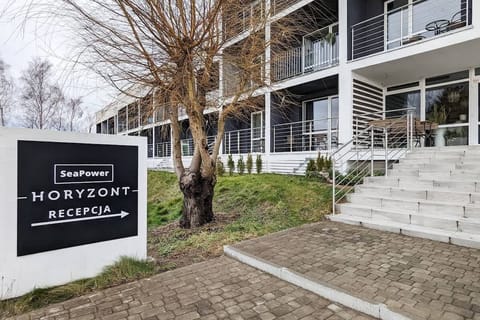 The exterior of the apartment building, featuring a landscaped entrance with stairs leading to the main door and a sign indicating the location.

