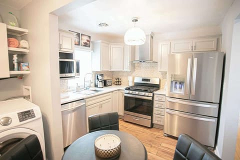 The kitchen, bathed in soft natural light, features contemporary stainless steel appliances, white cabinets, and a stone mosaic backsplash. A washer and dryer unit is integrated into the space, which opens to a cozy dining area with a round table.