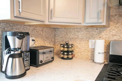 A modern kitchen corner boasts a marble countertop with a mosaic backsplash, housing a sleek coffee machine, toaster, paper towel holder, and a set of spice jars, all accentuated by cream-colored cabinetry.