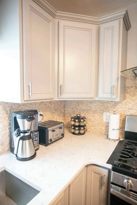 The elegant kitchen corner is well-appointed with modern appliances like a coffee machine and toaster, set against a granite countertop and a stone tile backsplash. Above, white cabinetry enhances the clean design, while natural light illuminates the spac