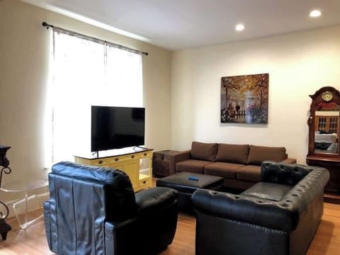The living room is comfortably furnished with a brown sectional sofa and a tufted leather ottoman. A large window draped with sheer curtains allows natural light to complement the warm tones of the room, which also features a classic wooden clock and a vi