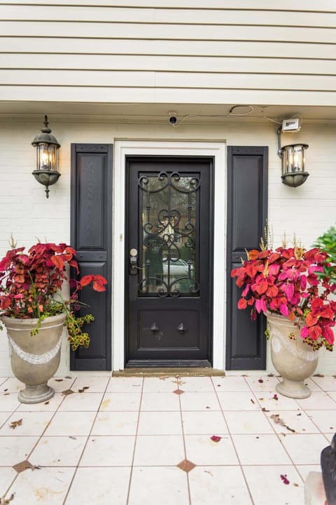 The image displays a welcoming front entryway with a classic black door featuring ornate glass detailing. Flanking the door are two large planters overflowing with vibrant red foliage, adding a pop of color to the neutral-toned façade.