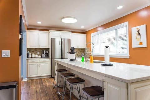 The image captures a modern kitchen with a warm ambiance, featuring terracotta walls and white cabinetry. A stainless steel refrigerator, contemporary appliances, and a kitchen bar with dark wooden stools accent the space.