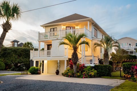A two-story house with a balcony, surrounded by palm trees and a garden. The exterior is lit with warm lights during dusk.
