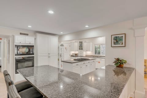 A modern kitchen with white cabinets, a built-in oven, a gas stove, and a large granite island. A framed picture hangs on the wall next to a small potted plant.