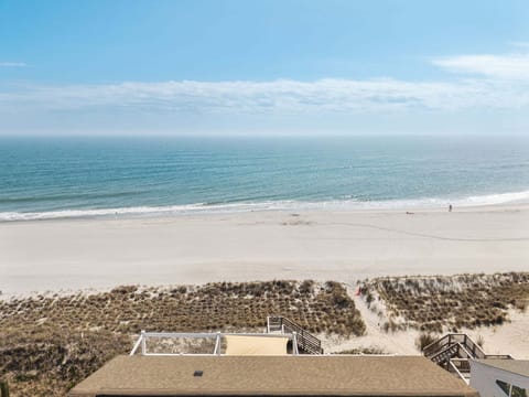 Aerial view of an oceanfront beach house marked near the shoreline, with neighboring coastal homes, wide sandy beach, rolling waves, and waterway views stretching along Oak Island.