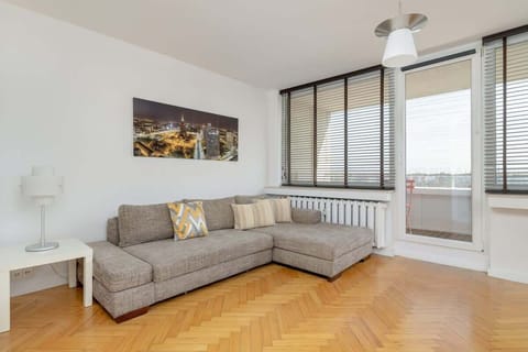 A bright living room with a large L-shaped sofa, wooden flooring, and panoramic windows offering a stunning city view.

