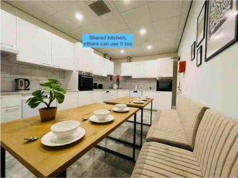 Shared kitchen with sleek white cabinets, modern appliances, a long wooden dining table, beige seating, a potted plant, and framed wall art for a cozy feel.