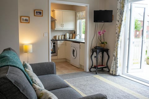 Lounge area leading to open kitchen. 2 seater sofa with cushions. TV on wall, small table