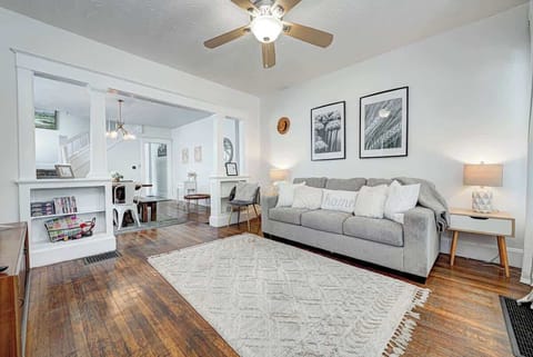 Cozy living room with a gray sofa, a soft textured rug, and wooden flooring, a ceiling fan and light fixture adding warmth. 