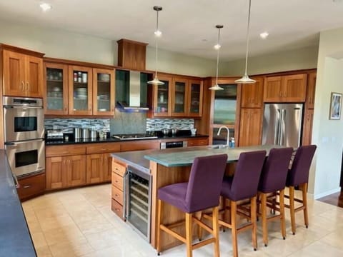 This expansive chef’s kitchen features custom wood cabinetry, a massive stainless steel island with a built-in prep sink, and elegant purple barstool seating.