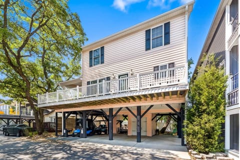 Raised light pink house with white railing balcony, black support beams, and shaded parking below.