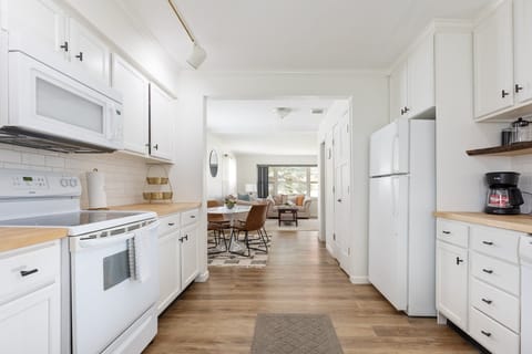 In our modern kitchen, white cabinets adorned with black handles stand tall alongside a warm wooden dining table, all illuminated by the soft glow of a radiant chandelier. It's a harmonious fusion of our cooking and dining realms, bathed in the gentle embrace of natural light, inviting creativity and connection into our home.