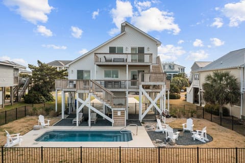 Rear view of a large elevated beach house with multiple decks, fenced yard, private pool, lounge chairs, and outdoor seating area.