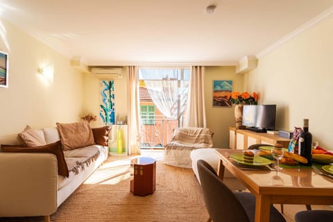 Bright living room with a comfortable sofa, TV, dining table, and window offering a view of the sunny outdoors. Perfect for relaxing.