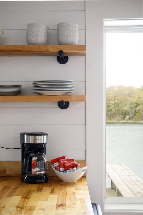 In the charming kitchen corner, wooden shelves display white dishware alongside a coffee maker, while a window provides a serene view of the tranquil lake.