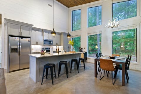 A modern kitchen and dining area with large windows, featuring an island with bar stools, stainless steel appliances, white cabinets, and the dining area to the right.