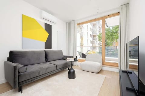 A modern living room with a grey sofa, black furniture, and a yellow geometric wall art piece, illuminated by natural light from large windows.