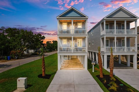 Stunning coastal rental featuring elegant architecture with wrap-around porches and palm-lined entrance under brilliant sunset skies.