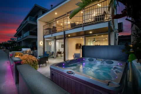 A luxurious two-story beachfront home illuminated at sunset, featuring expansive glass windows, multiple outdoor seating areas, and a shared hot tub. This stunning property offers a perfect blend of relaxation and sophistication with breathtaking views of