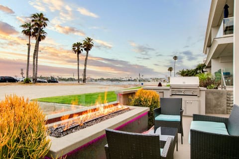 A cozy beachfront patio featuring a modern fire pit, comfortable wicker seating with blue cushions, and seamless access to the indoor living space. Surrounded by lush tropical plants, this inviting outdoor area is perfect for relaxing and enjoying warm ev