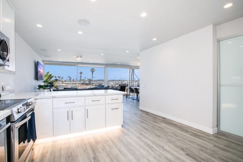 A bright and spacious open-plan living area featuring a modern kitchen with sleek white cabinetry, stainless steel appliances, and under-cabinet lighting. The room seamlessly flows into a comfortable seating area with large windows offering breathtaking v