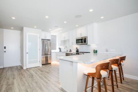 A sleek and modern kitchen featuring white cabinetry, stainless steel appliances, and a large marble island with stylish wooden bar stools. The open layout and ample lighting create a bright and inviting space perfect for cooking and casual dining.