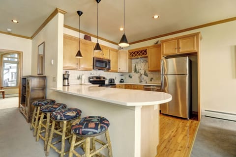 A modern kitchen with light wood cabinetry, stainless steel appliances, and a curved countertop with four patterned stools. There are hanging pendant lights above the counter and a backsplash featuring a scenic design.