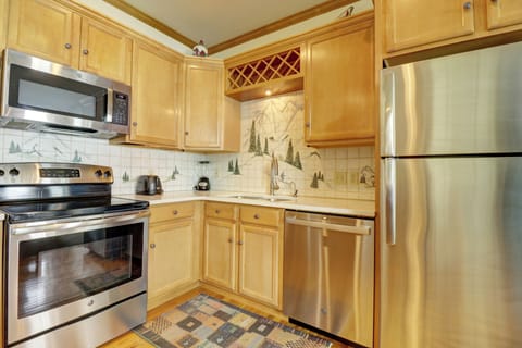 A modern kitchen with stainless steel appliances, including a microwave, stove, dishwasher, and refrigerator. Wooden cabinets and a tiled backsplash with tree designs complete the look.