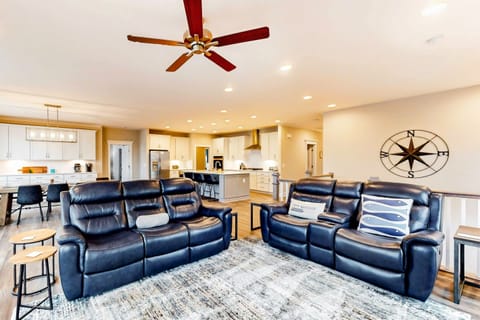 A modern living room with plush leather recliners, a ceiling fan above, opens into a bright kitchen with white cabinetry and bar-style seating