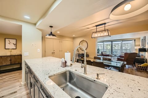 Modern kitchen with a marble countertop island, built-in sink, and stainless steel fixtures. In the background, there is a dining area with a wooden table, chairs, and a well-lit living space.